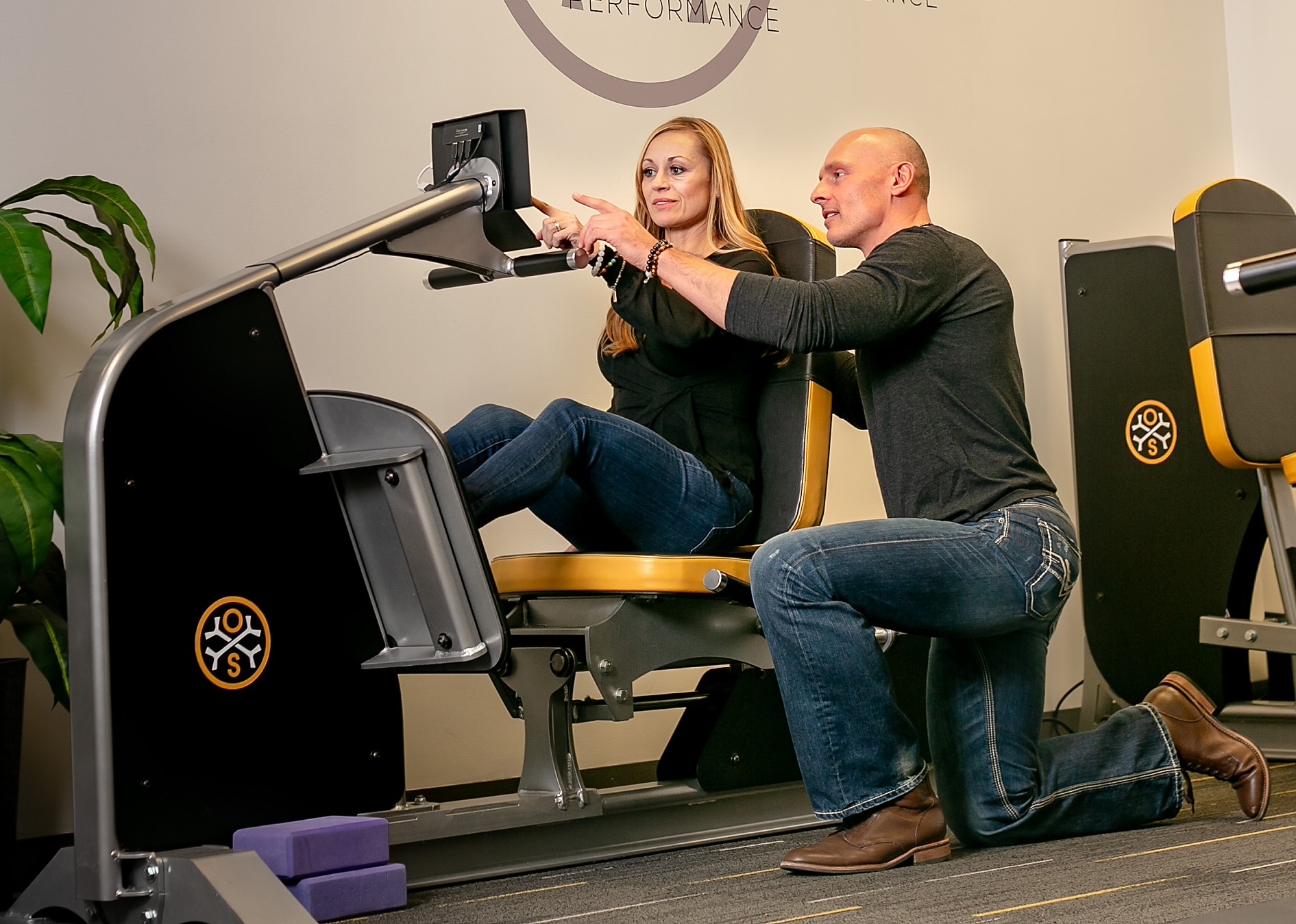 An OsteoStrong coach kneeling beside a member, guiding her through a session on the Spectrum device